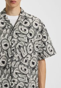 Camicia a maniche corte in bianco e nero, caratterizzata da un motivo astratto lucido con lettere ingrandite. Bottoni lungo la patta e texture del tessuto morbido.
