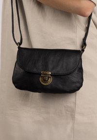 Borsa a tracolla nera in pelle con un flap arrotondato, chiusura in ottone e tracolla sottile. Texture liscia con dettagli di cucitura sottili.