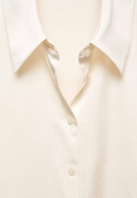 Blouse blanche en soie avec un col classique et trois boutons visibles semblables à des perles le long de la patte de boutonnage avant.