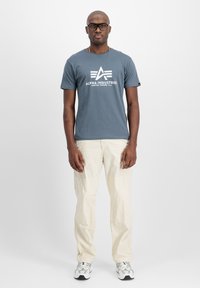 Blaues Baumwoll-T-Shirt mit weißem Drucklogo, kombiniert mit hellbeigen Cargo-Hosen. Das Modell trägt außerdem silberne Sportschuhe und schwarze Brille.