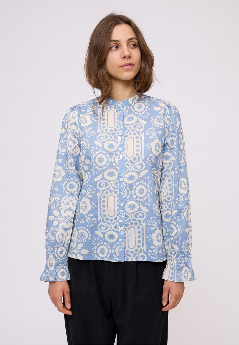 Camicia azzurra a maniche lunghe con motivi floreali e geometrici bianchi, con chiusura a bottoni e polsini arricciati. Tessuto morbido con una vestibilità rilassata.