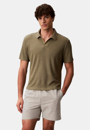 Ung mand med kort krøllet hår iført en tekstureret olivengrøn kortærmet polo og lyse stribede shorts, hænderne i lommerne.