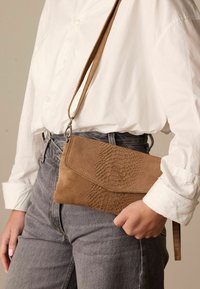 Bruine leren clutch met een geperst reptielpatroon, voorzien van een afneembare strap en een klep sluiting, gehouden tegen grijze spijkerbroeken.