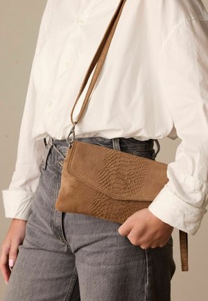 Brown leather clutch with embossed reptile pattern, featuring a detachable strap and a flap closure, held against gray jeans.