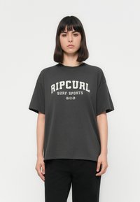 Jaunas žmogus su tiesiais tamsiais plaukais, vilkintis laisvą tamsiai pilką "Ripcurl Surf Sports" marškinėlį ir juodas kelnes, stovi priešais baltą foną.