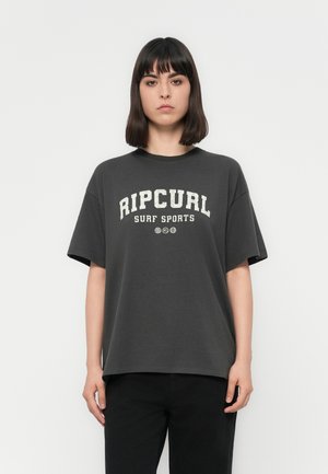 Young person with straight dark hair wearing a loose dark gray Ripcurl Surf Sports t-shirt and black pants, standing against white background.