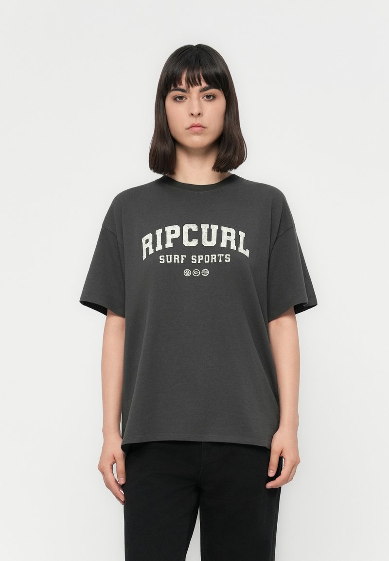 Jaunas žmogus su tiesiais tamsiais plaukais, vilkintis laisvą tamsiai pilką "Ripcurl Surf Sports" marškinėlį ir juodas kelnes, stovi priešais baltą foną.