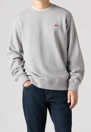 Homme portant un sweat-shirt à col rond Levi’s gris clair et un jean bleu foncé, debout avec une main dans la poche sur un fond uni.