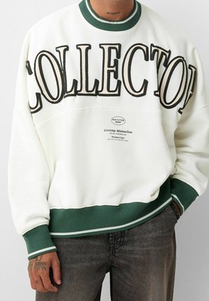 Person trägt ein weißes Sweatshirt mit grünen, gerippten Bündchen und Saum, großem „COLLECTOR“-Schriftzug und kleinerem aufgedrucktem Text auf der Brust, sichtbare Handtätowierung.