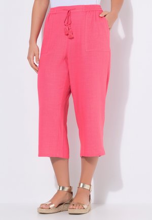 Mujer con pantalones cortos fucsia brillantes con bolsillos frontales y cintura con cordón, combinados con sandalias doradas con pulsera en el tobillo.