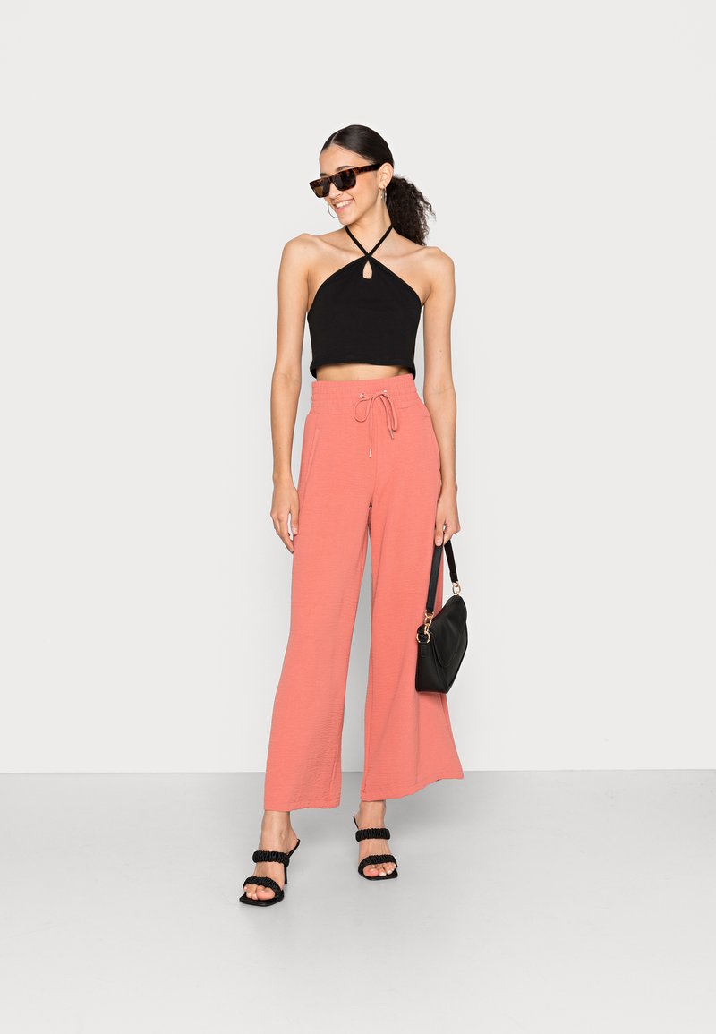 Top cropped preto com alças, calças largas rosas com cintura com cordão, mala preta e sandálias pretas com tiras. Óculos de sol completam o visual.