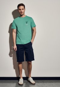 Mátově zelené bavlněné tričko s grafikou humra, spárované s tmavě námořnickými cargo šortkami. Nosené s krémovými sneakers. Jednoduché pozadí.