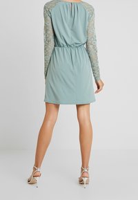 Robe bleu-vert clair avec de longues manches en dentelle, taille élastique et détail en forme de clé à l'arrière. Texture de tissu lisse et design à hauteur des genoux. Talons hauts argentés.