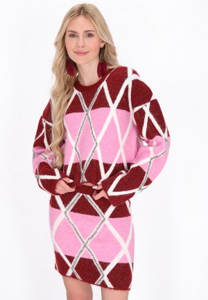  Blond kvinde iført en rød og lyserød argyle mønstret sweaterkjole og røde fjerøreringe står mod en hvid baggrund.