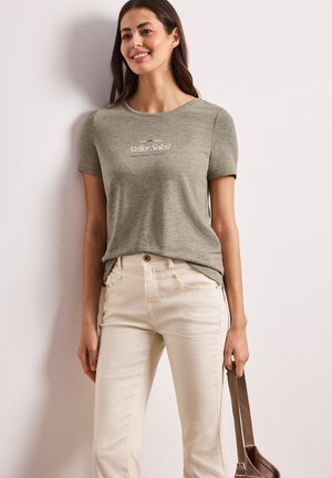Femme souriante portant un t-shirt vert olive "Atelier Soleil" et un pantalon crème, tenant un sac à bandoulière en cuir marron, debout contre un mur blanc.