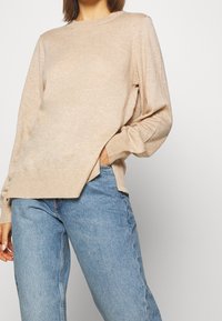 Pull beige en maille avec un col rond, des poignets côtelés et une fente latérale. Assorti à un jean en denim bleu clair. Texture douce, coupe décontractée.