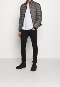 Grå puffjakke med lynlås foran, stående krave og kontrasterende blå foring, parret med en hvid t-shirt og sorte bukser. Sorte støvler.