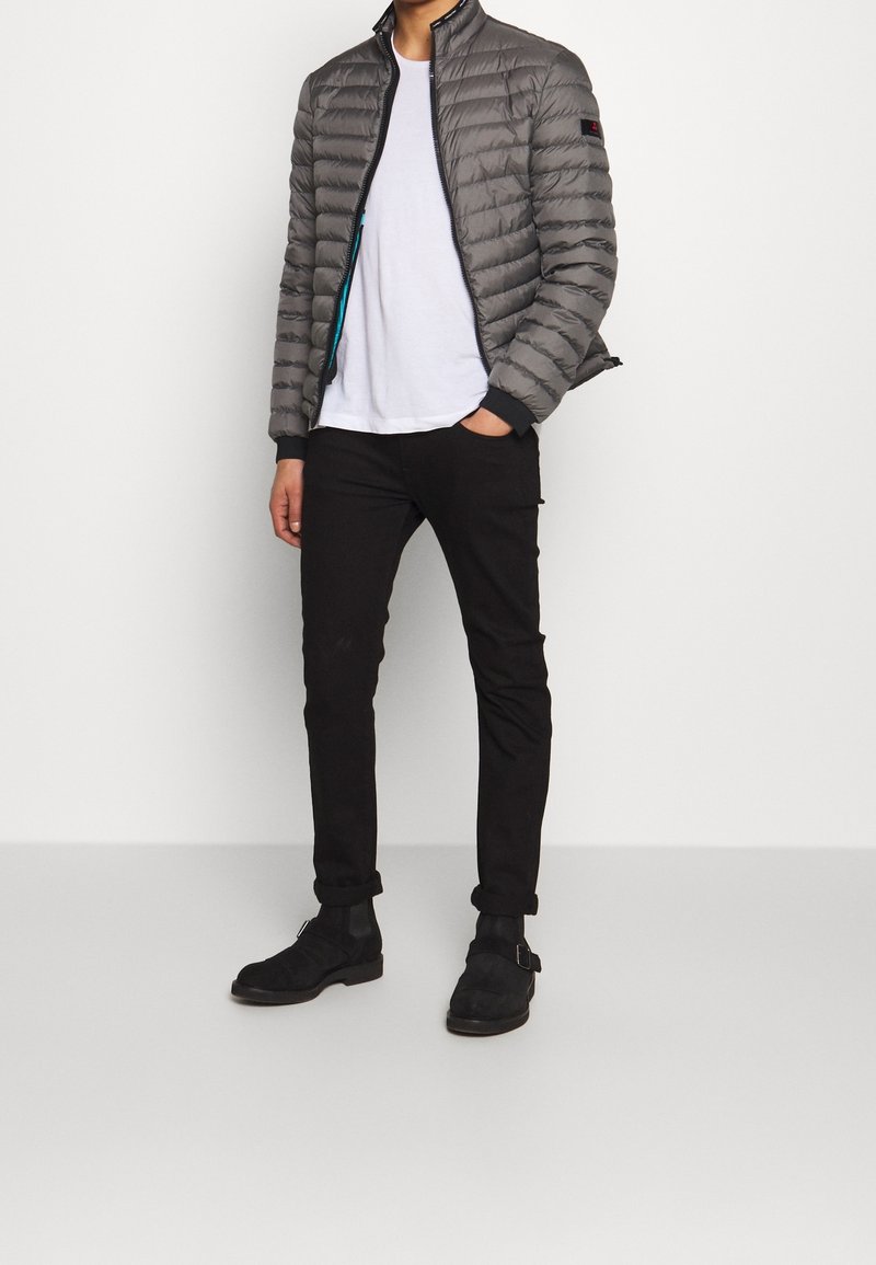 Grå puffjakke med lynlås foran, stående krave og kontrasterende blå foring, parret med en hvid t-shirt og sorte bukser. Sorte støvler.