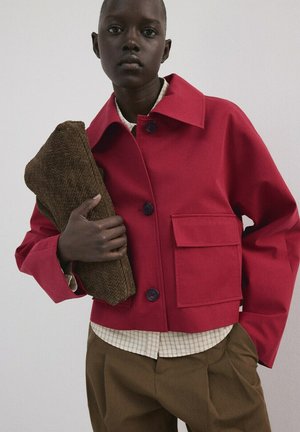 Mannequin portant une veste rouge boutonnée avec une grande poche, une chemise beige à carreaux, un pantalon marron, tenant une pochette marron texturée.