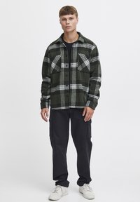 Cămașă din flanel cu carouri verzi și albe, cu două buzunare pe piept; purtată peste un top negru, asortată cu pantaloni cargo negri și adidași albi.