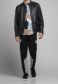 Zwarte leren bomberjack over een meerkleurrig geometrisch shirt, gecombineerd met zwarte cargobroeken met grijze accenten en zwarte sneakers.