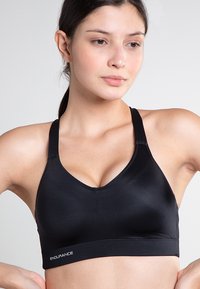 Svart sport-bh med en djup V-urringning, tillverkad av mjukt och elastiskt material. Har breda axelremmar och en logotyp på bandet nedanför.