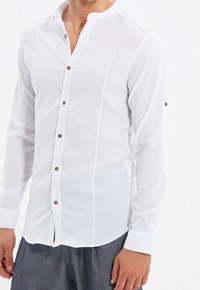Biała koszula na długi rękaw, zapinana na guziki, z kołnierzem typu mandarynka, wykonana z teksturowanej tkaniny, z brązowymi guzikami i dopasowanym krojem, z rozcięciami po bokach u dołu.