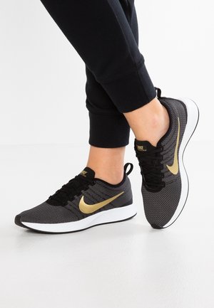 Stopy w czarno-złotych butach do biegania Nike z białymi podeszwami, w połączeniu z czarnymi spodenkami dresowymi, na białym tle podłogi.