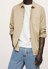 Chemise beige à boutons avec manches longues et une poche poitrine, portée sur un t-shirt blanc à col rond, associée à un jean bleu clair.