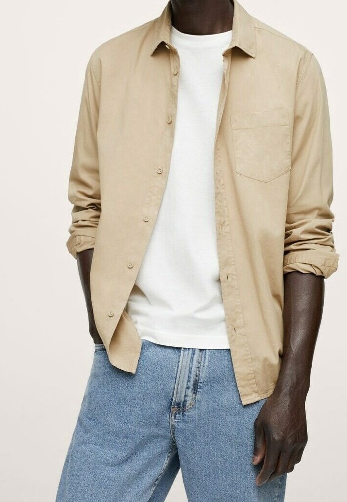 Chemise beige à boutons avec manches longues et une poche poitrine, portée sur un t-shirt blanc à col rond, associée à un jean bleu clair.