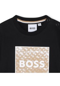 Crna pamučna majica s okruglim izrezom, s bež grafičkim kvadratnim dizajnom i riječi "BOSS" u bijeloj boji. Akcenti s gumbima na vratu.