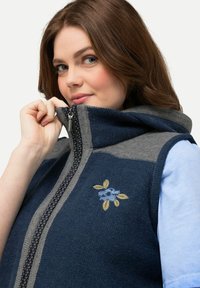 Námorně modrá vesta s šedými akcenty, vyrobená z texturované tkaniny. Obsahuje květinový vyšívací design a zapínání na zip. Styl s kapucí.