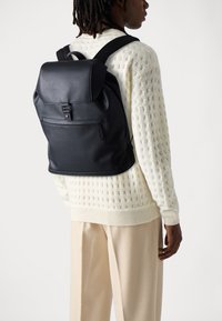 Black leather backpack with a flap closure, adjustable straps, and front zip pocket. Worn over a textured white sweater and beige trousers.