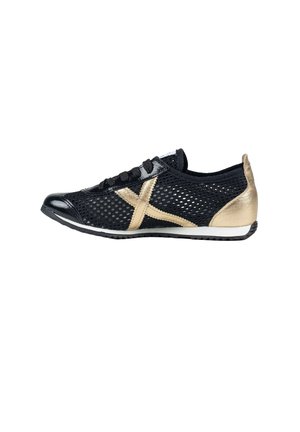 Zapatilla deportiva de malla negra con logo X dorado y parche en el talón, cordones negros y suela de perfil bajo, vista de lado sobre fondo blanco.