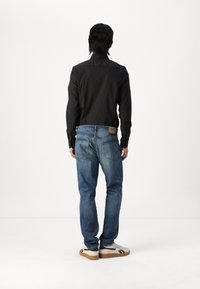 Camisa negra de manga larga, jeans de mezclilla azul con bolsillos traseros y zapatillas blancas con detalles en negro. Diseño sencillo, apariencia casual.