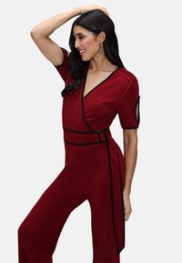 Tutina rossa con design avvolgente, bordi neri, maniche corte e una cintura in evidenza. Tessuto liscio e taglio ampio.