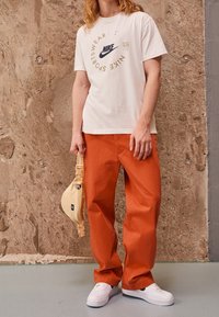 T-shirt blanche avec logo Nike noir et texte, pantalon orange, baskets blanches, et sac à épaule jaune clair, se tenant sur une surface grise texturée.