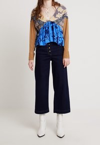 Pestrobarevná blůza s květinovými a vzorovanými detaily, s volánkovým modrým dolním dílem; v kombinaci s tmavými džíny s vysokým pasem a bílými kotníkovými botami.