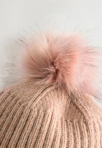 Pehmest beežist lõngast kootud beanie, millel on vertikaalne ribakudu, ja millel on pehme roosa pom-pom tipus. Materjal näeb välja soe ja tekstureeritud.