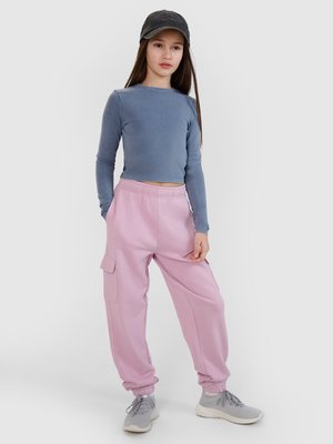 Młoda dziewczyna nosząca czarną czapkę, niebieski crop top z długim rękawem, różowe spodnie cargo oraz szare trampki, stojąca na jednolitym białym tle.