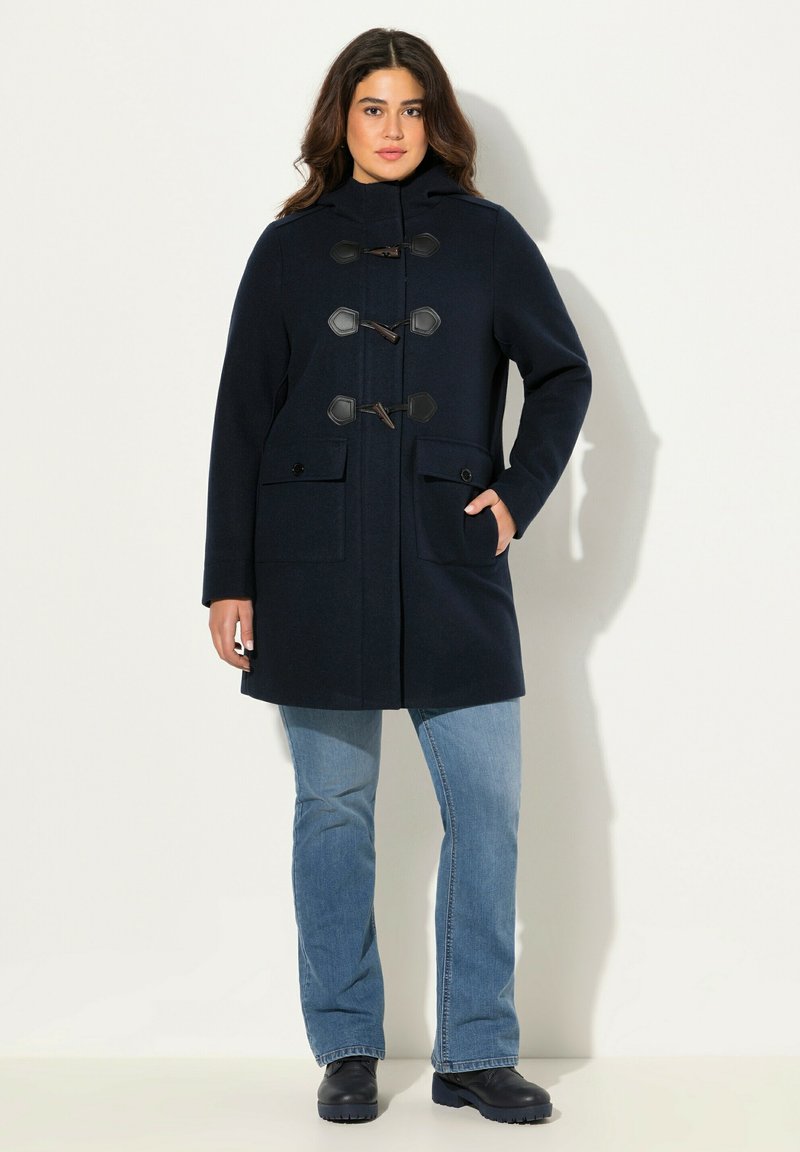 Cappotto di lana blu navy con cappuccio, chiusure a fibbia e tasche laterali. Abbinato a jeans blu chiaro e scarpe nere.