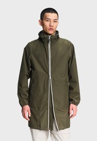 Jeune homme portant un long imperméable vert olive avec une fermeture éclair à l'avant, des poignets élastiques et des poches latérales zippées, se tenant devant un fond clair.