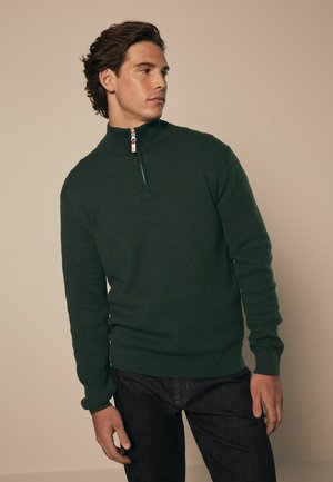 Jersey de punto verde con cuello alto, media cremallera, mangas largas y puños y bajo acanalados. Combinado con jeans de mezclilla oscura.