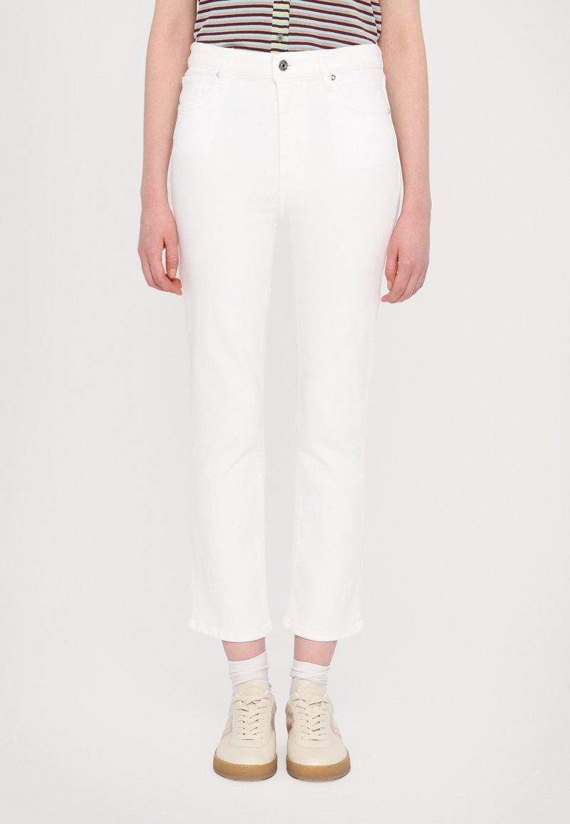 Personne portant un pantalon blanc court, des baskets beige clair avec des chaussettes blanches, et une chemise rayée avec des boutons visibles.