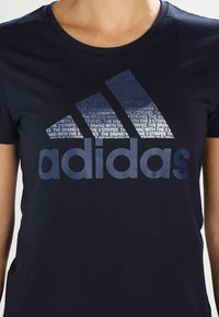 Námořnická modrá bavlněná tričko s velkým logem Adidas v tmavě modré barvě s textovým vzorem v pozadí. Klasický design s kulatým výstřihem.