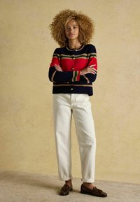 Navy Cardigan mit roten und grünen Streifen, goldenen Knöpfen. Weiße hochtaillierte Hose und braune Slipper runden das Outfit ab. Texturierten Hintergrund.