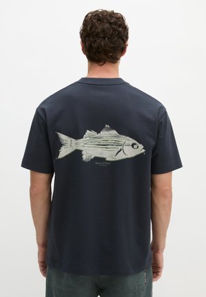 Mężczyzna odwrócony tyłem, ubrany w granatowy t-shirt z dużą jasnoszarą grafiką ryby na plecach.