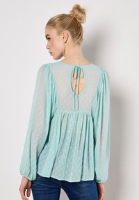 Helle blaue Bluse mit langen, puffigen Ärmeln, strukturiertem Stoff, Schlüsselrücken mit Bindeschluss und gerafftem Saum für eine fließende Silhouette.