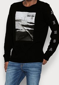 Mustapaita pitkällä hihalla, jossa keskellä on grafiikka tie-tunnelmasta. "EAGLE USA" painettuna hihoihin valkoisella. Pehmeää puuvillakangasta.