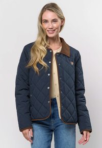 Jonge vrouw met lang blond haar, gekleed in een marineblauwe doorgestikte jas met bruine bies, een beige blouse met knopen en een blauwe spijkerbroek, glimlachend naar de camera.